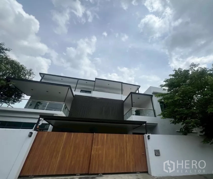Home Office for rent in Watthana, Bangkok - Modern multi‑level home office exterior with white walls, glass balconies and a wide wooden gate under a cloudy sky.