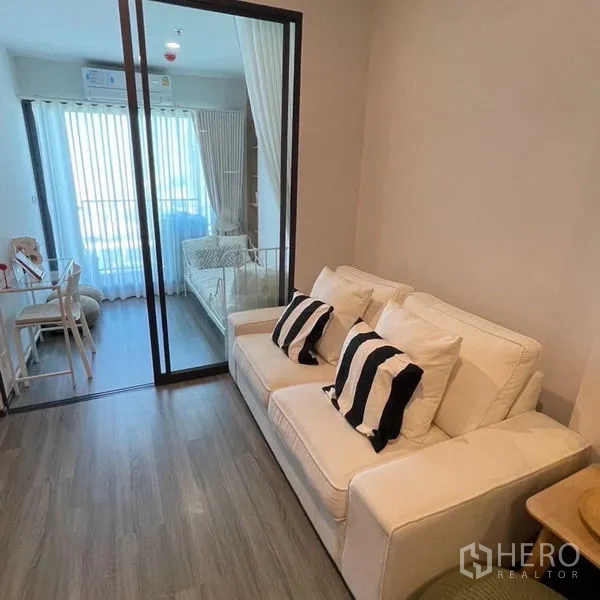 คอนโด ให้เช่า - ห้องนั่งเล่นสว่าง โซฟาสีขาว พื้นไม้ และประตูกระจกบานเลื่อนเชื่อมสู่ห้องนอนและระเบียง