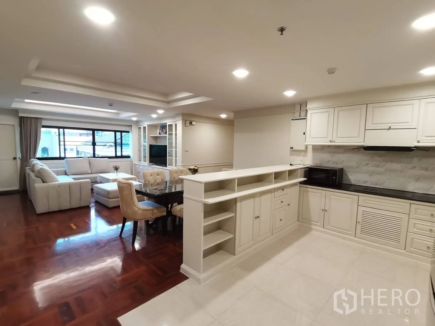 Condominium for rent in Watthana, Bangkok - Open-plan living room with L-shaped sofa, dining set, and white kitchen with recessed lighting and wood floors.