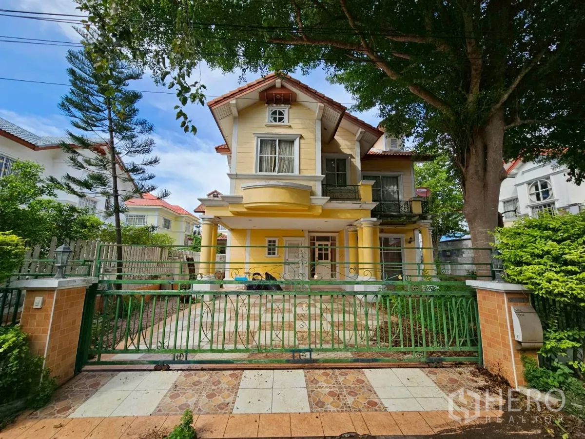 Detached House for rent or sale in Bang Lamung, Chon Buri - Front view of a two-storey yellow detached house with red-tile roof, green gate, and a shaded driveway.