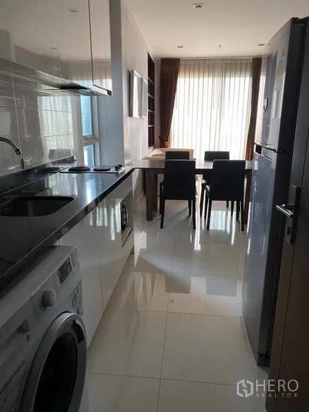 Condominium for rent in Watthana, Bangkok - Galley kitchen with glossy white cabinets, black countertop, sink, cooktop, fridge and washing machine leading to dining area