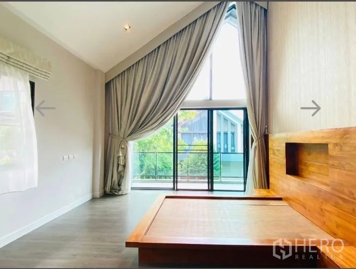 Detached House for sale in Saphan Sung, Bangkok - Master bedroom with high ceiling, tall windows and a wooden platform bed opening to a balcony.