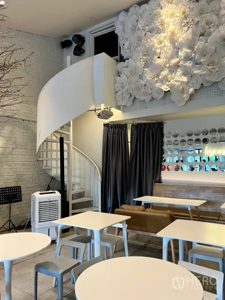 零售空间 出租在Watthana, Bangkok - 明亮的零售咖啡空间，白色螺旋楼梯、砖纹墙面与花艺背景墙，配现代桌椅。