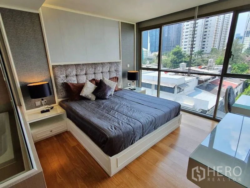 Condominium for rent in Watthana, Bangkok - Corner bedroom with large windows, upholstered headboard, bedside lamps, and wood flooring with city outlook.
