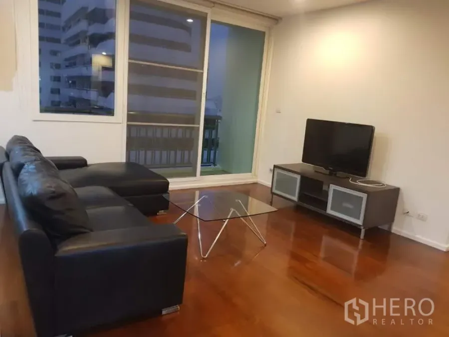 Condominium for rent in Watthana, Bangkok - Bright living room with black leather sectional, glass coffee table, TV console, wood floors, and sliding doors to a balcony with city views.