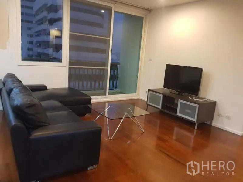 Condominium for rent in Watthana, Bangkok - Bright living room with black leather sectional, glass coffee table, TV console, wood floors, and sliding doors to a balcony with city views.