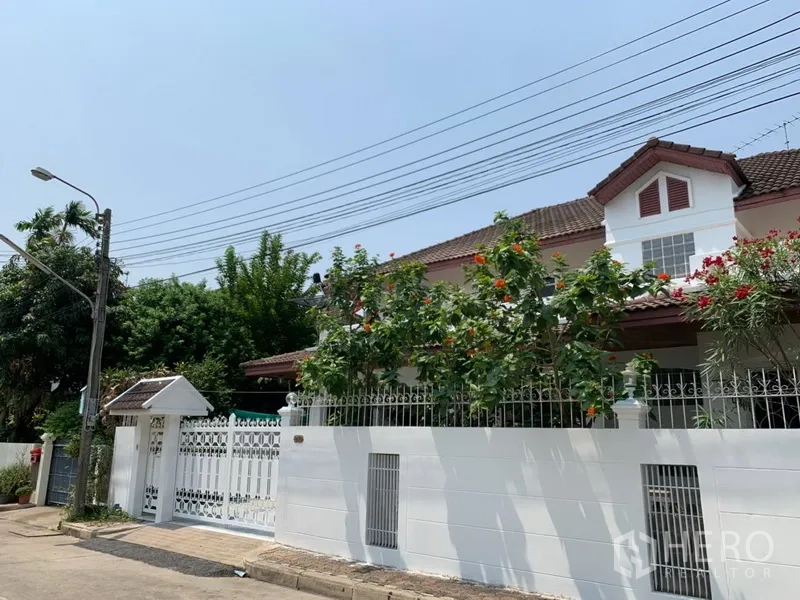 Detached House for rent in Khlong Toei, Bangkok - Two-storey detached house with white perimeter wall, gated entrance and red-tiled roof on a quiet street with flowering trees.