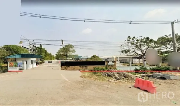 工厂 出售在Lam Luk Ka, Pathum Thani - 林路甲工业园入口，设有门岗、围墙与宽阔的进出道路，架空电线可见。