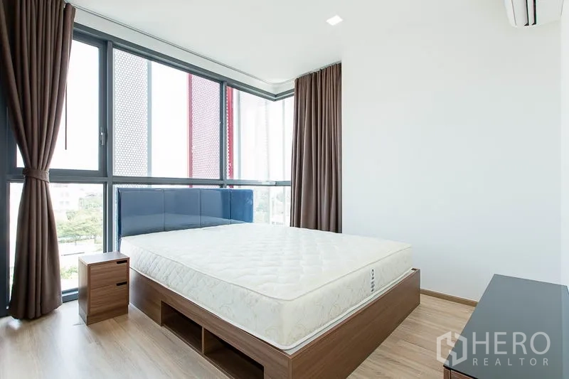 Condominium for rent in Watthana, Bangkok - Bedroom with wide windows on two sides, brown curtains and wall-mounted air conditioner.