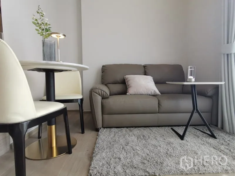 公寓 出租 - 舒适客厅，灰色沙发、圆形餐桌与边几，脚踏地毯，旁有落地窗与窗帘。