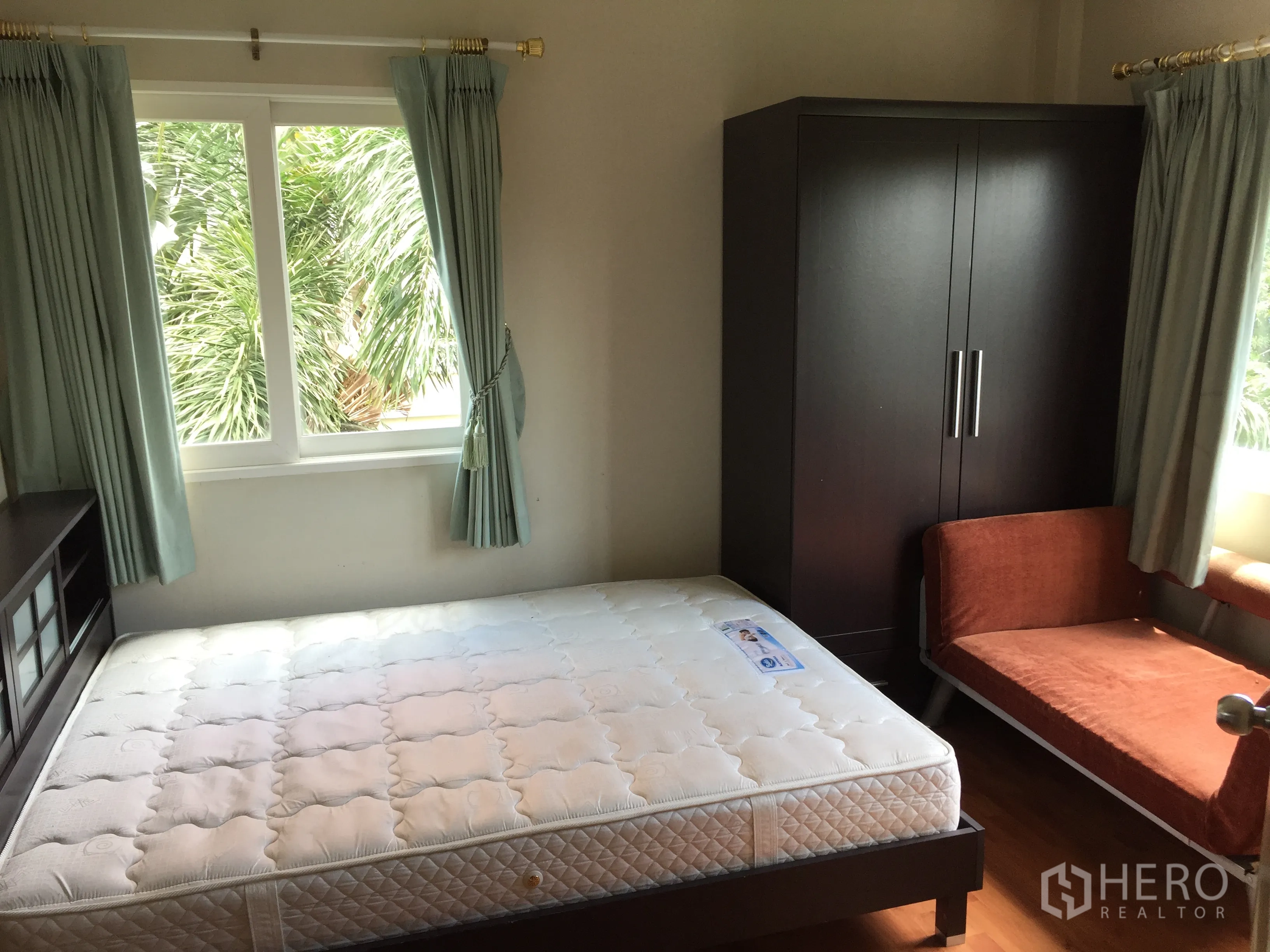 Townhouse for rent or sale in Bueng Kum, Bangkok - Bright bedroom with large windows, green curtains, bed and dark wardrobe overlooking lush trees.