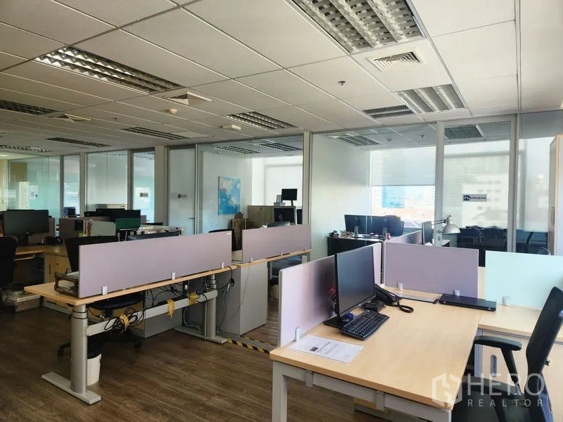 Office Space for rent - Open workspace with desks and monitors beside glass-partitioned meeting rooms and ceiling lights.