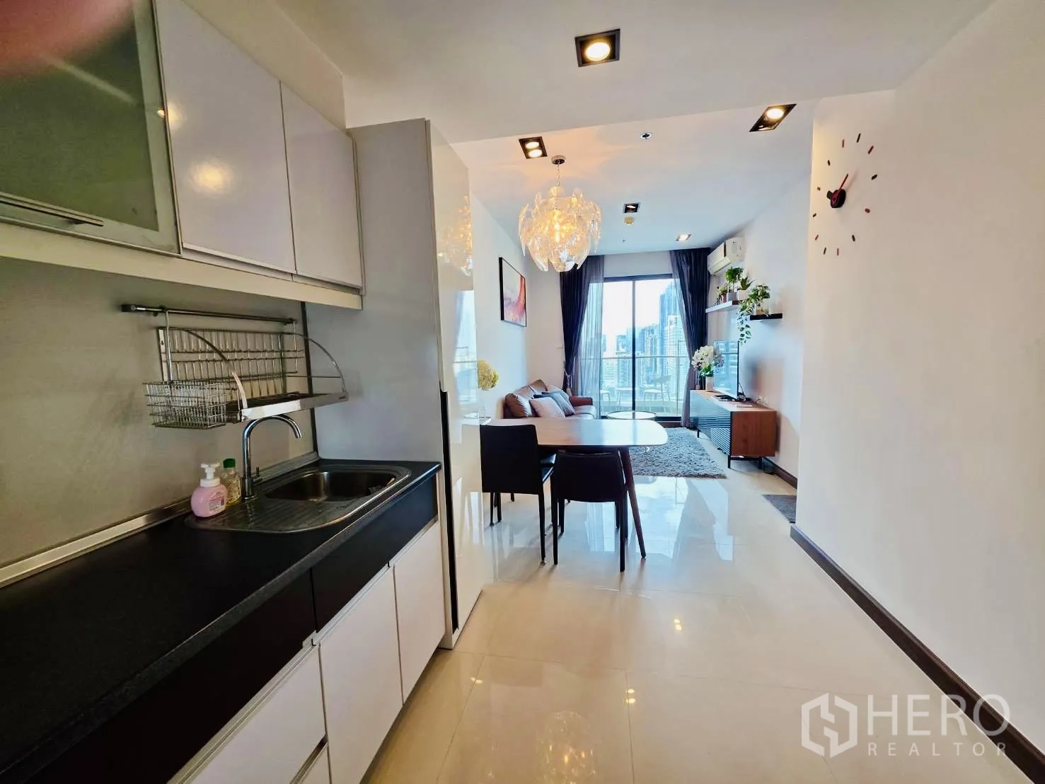 Condominium for rent in Huai Khwang, Bangkok - Galley kitchen with white cabinets and sink opening to dining and bright living room with balcony view.