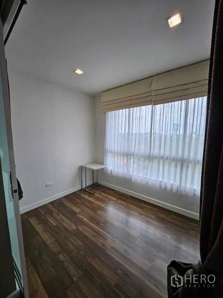 Condominium for sale in Watthana, Bangkok - Compact room with large window, sheer curtains, and wooden flooring ideal for study or second bedroom.