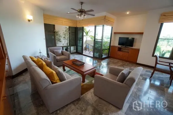 อพาร์ทเมนท์ ให้เช่าในWatthana, Bangkok - มุมนั่งเล่นโซฟา พัดลมเพดาน และประตูกระจกเปิดสู่ระเบียงมีต้นไม้