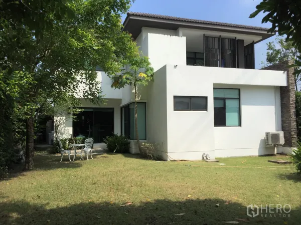 Detached House for rent or sale in Prawet, Bangkok - Shaded back garden with lawn, mature trees and a patio set beside the white facade and sliding glass doors.