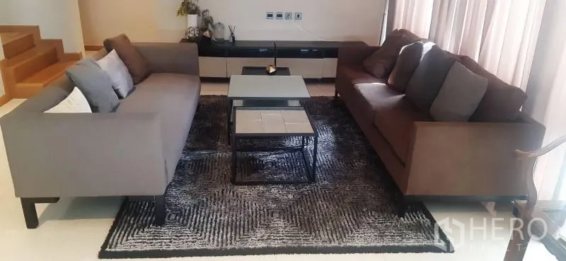 Detached House for rent or sale in Huai Khwang, Bangkok - Modern living room with two grey sofas, nesting coffee tables and a textured rug beside a wooden staircase.