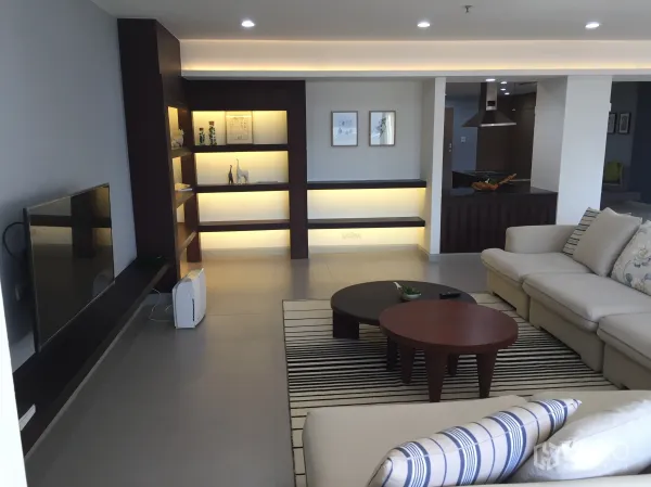 Condominium for sale in Watthana, Bangkok - Spacious living room with backlit built-in shelves, sectional sofa, round coffee tables, TV wall and recessed lighting beside an open kitchen.