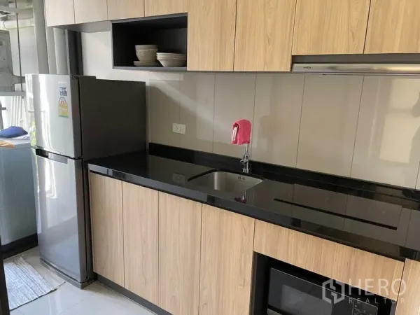 Condominium for sale in Watthana, Bangkok - Modern kitchenette with wood cabinets, black stone countertop, sink, and refrigerator.