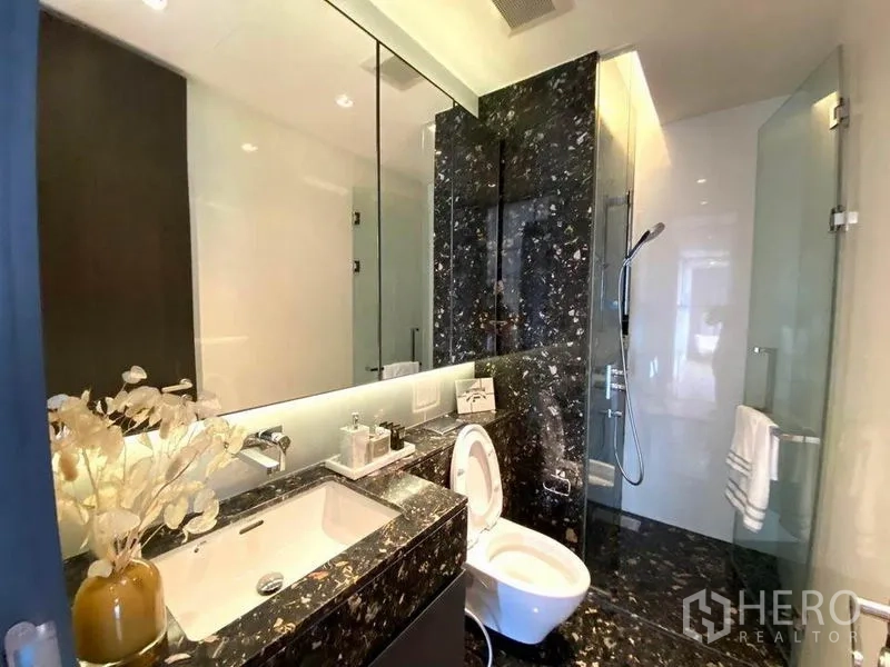 Condominium for rent or sale in Khlong Toei, Bangkok - Modern bathroom with stone vanity, glass shower enclosure and backlit mirror.