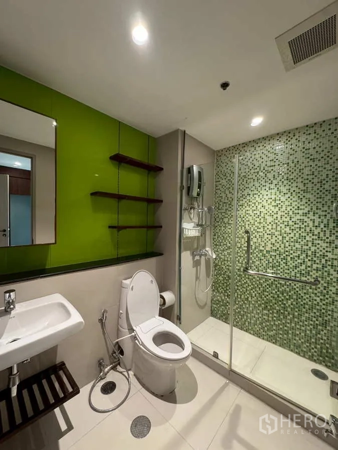 Condominium for rent or sale in Khlong Toei, Bangkok - Bathroom with white fixtures, green accent wall and glass shower enclosure.