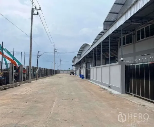Warehouse for rent in Krathum Baen, Samut Sakhon - Modern warehouse row with curved metal roofs along a wide concrete access road and secure perimeter fencing in Krathum Baen.