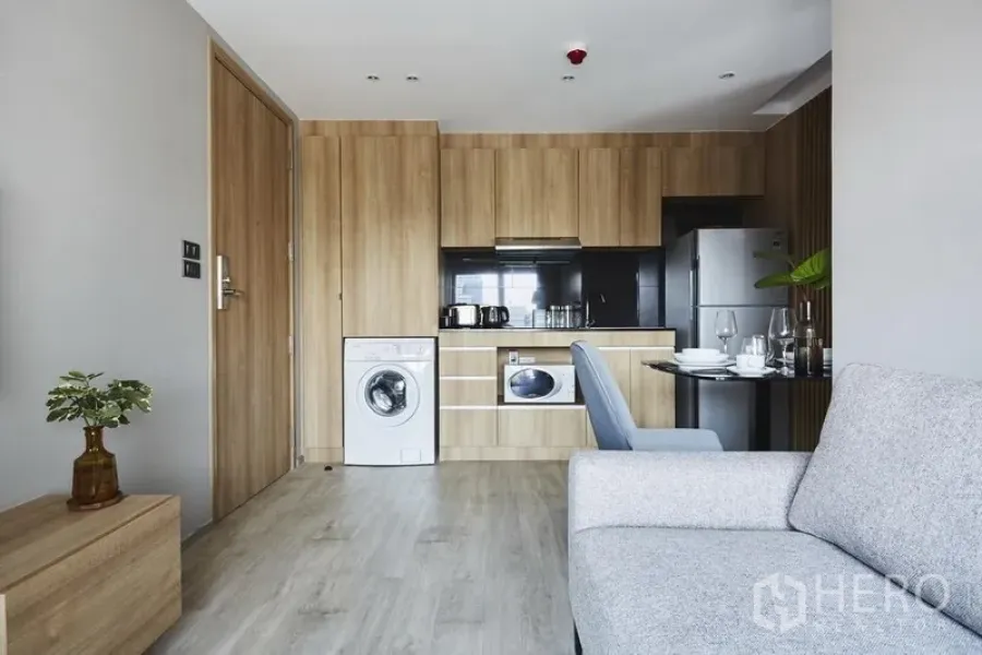 Apartment for rent in Khlong Toei, Bangkok - Open-plan kitchen with wood cabinetry, fridge, washing machine and small dining nook.