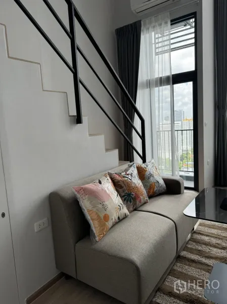 Condominium for rent in Huai Khwang, Bangkok - Cozy sofa beneath a staircase with city views through large windows and sheer curtains.