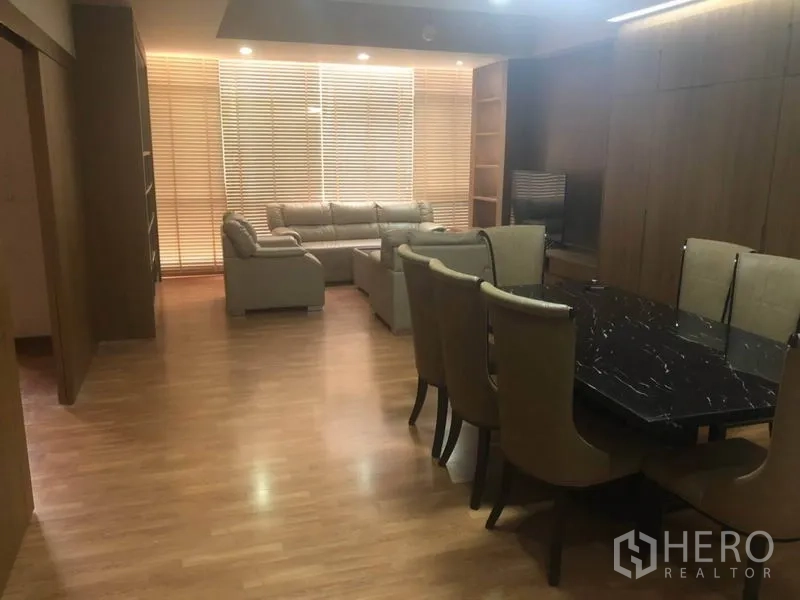 Condominium for sale in Bang Na, Bangkok - Open-plan living and dining area with wood floors, black marble dining table, beige sofas, TV unit and large window blinds.