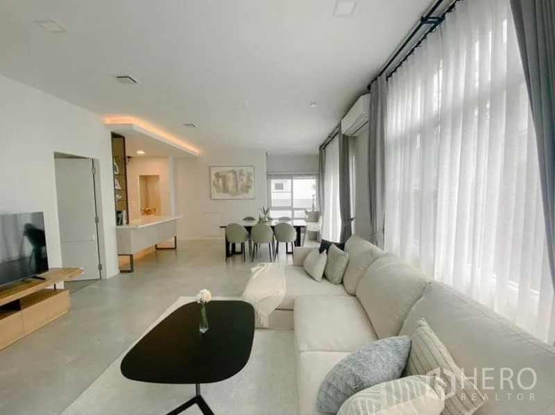 Detached House for rent in Prawet, Bangkok - Living room with floor-to-ceiling sheer curtains, a TV console and dining set beyond.