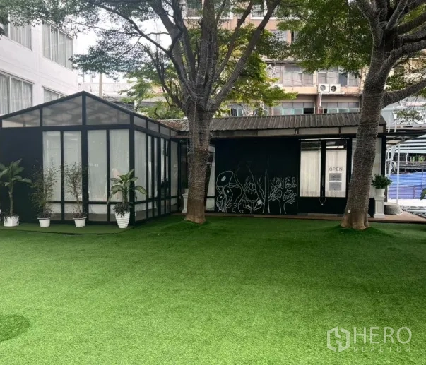 零售空间 出租在Watthana, Bangkok - 宽阔草坪外摆区，两棵大树与相连的玻璃亭式商铺外观。