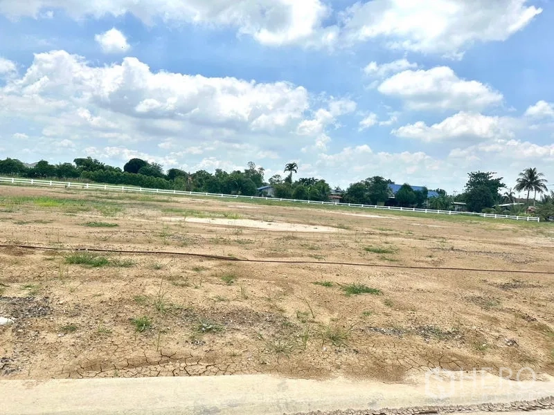 Land for sale in Lam Luk Ka, Pathum Thani - Flat bare soil plot with sparse grass and rural homes beyond a white rail fence.