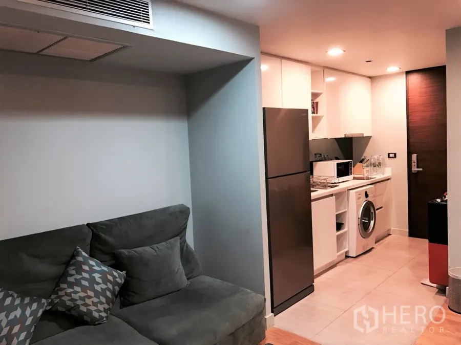 Condominium for rent or sale in Bang Rak, Bangkok - Open-plan living room with gray sofa and a compact white kitchen featuring a fridge and in-unit washer in a Silom condo.