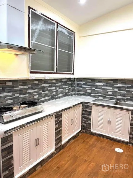 联排别墅 出租在Bang Na, Bangkok - L形厨房带水槽、白色柜体、灰色马赛克挡板与仿木地面。