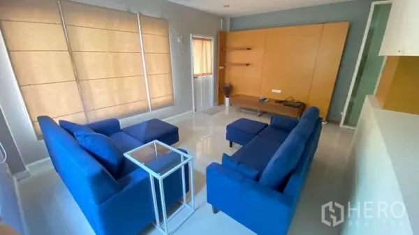 บ้าน ให้เช่า - ห้องนั่งเล่นอบอุ่น โซฟาสีน้ำเงินเข้าชุด ผนังวางทีวีพร้อมชั้นวาง และมู่ลี่เต็มบาน