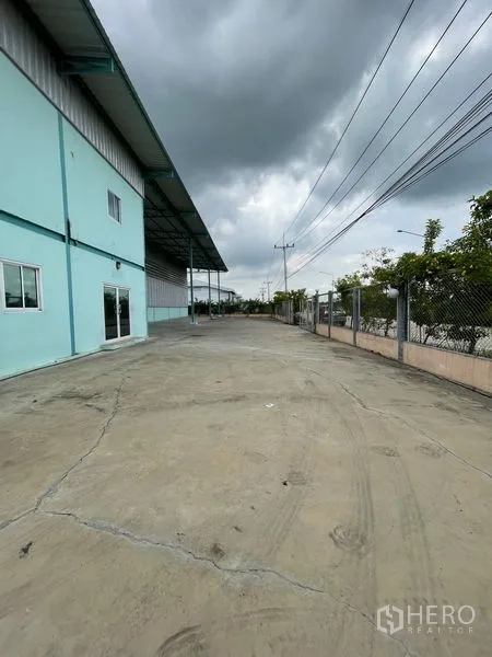 Warehouse for rent in Lat Lum Kaeo, Pathum Thani - Exterior driveway beside the warehouse with covered loading edge, perimeter fencing and power lines under a cloudy sky.