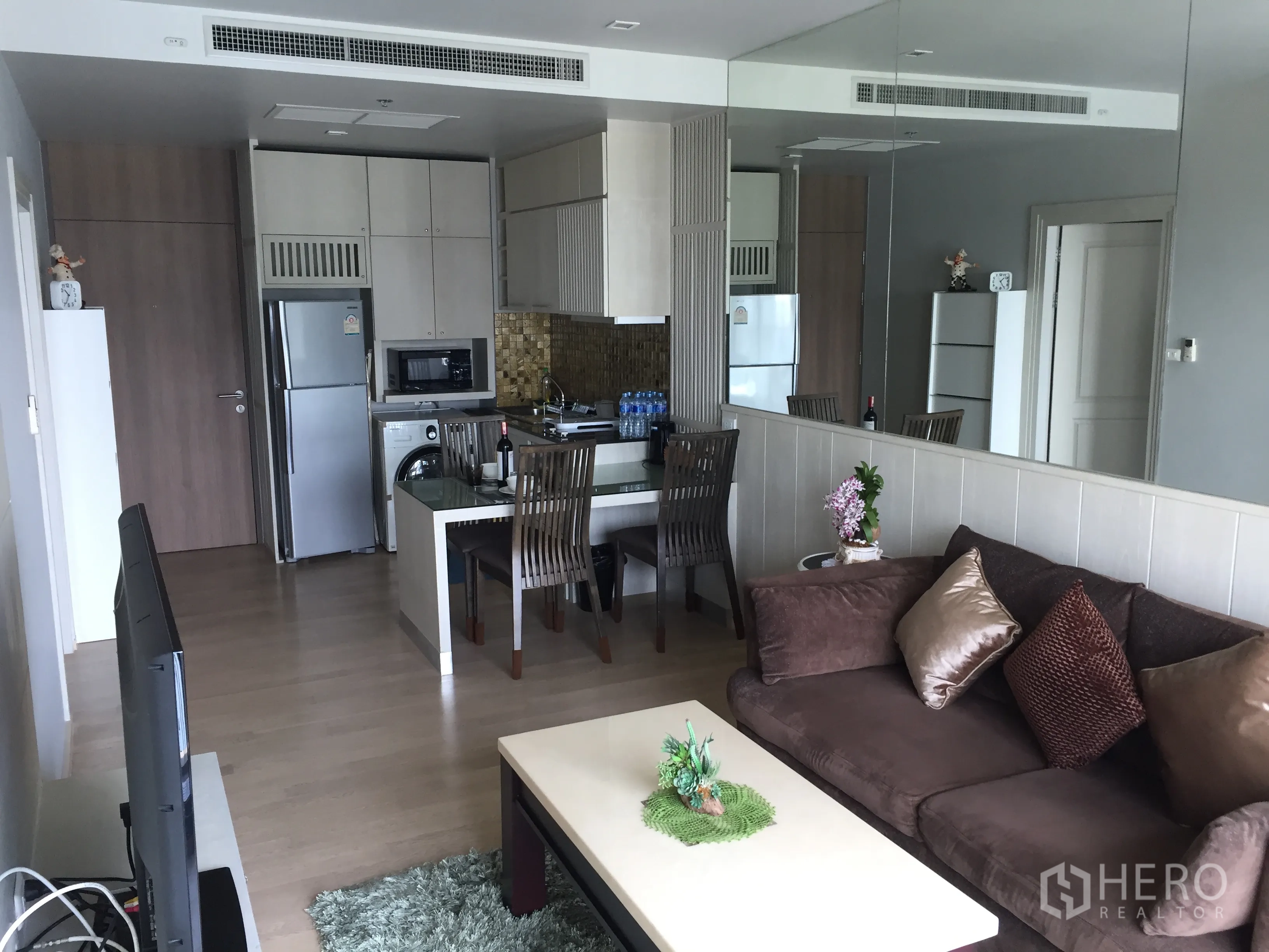 公寓 出租在Bangkok - 开放式客厅带镜面墙，沙发、电视与餐区相连的小巧厨房。