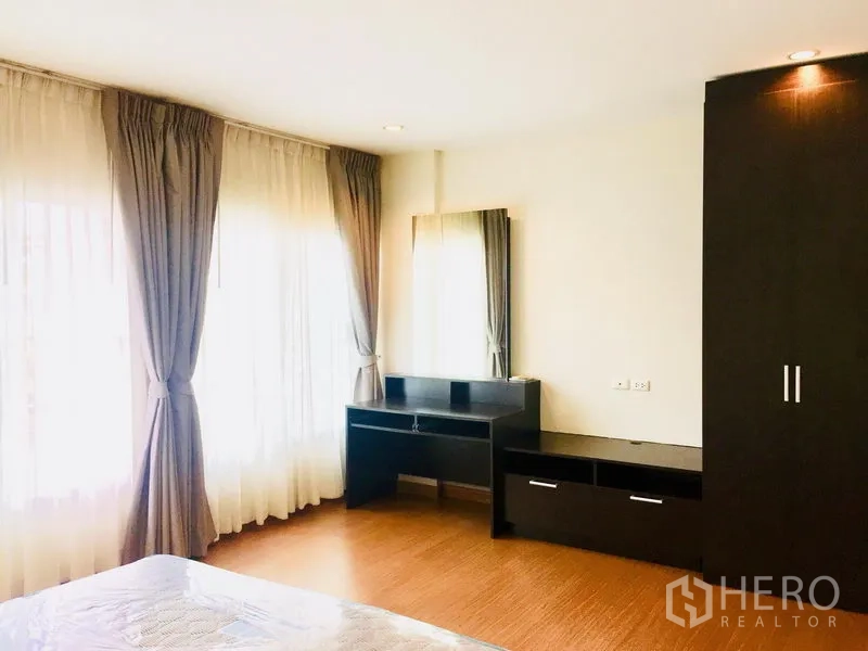 Townhouse for rent in Prawet, Bangkok - Spacious bedroom with corner windows, dark built-in cabinet, and dressing desk.