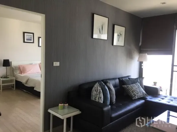 Condominium for rent in Watthana, Bangkok - Modern living room with black L-shaped sofa, gray feature wall, doorway to separate bedroom, and bright window with roller blinds.