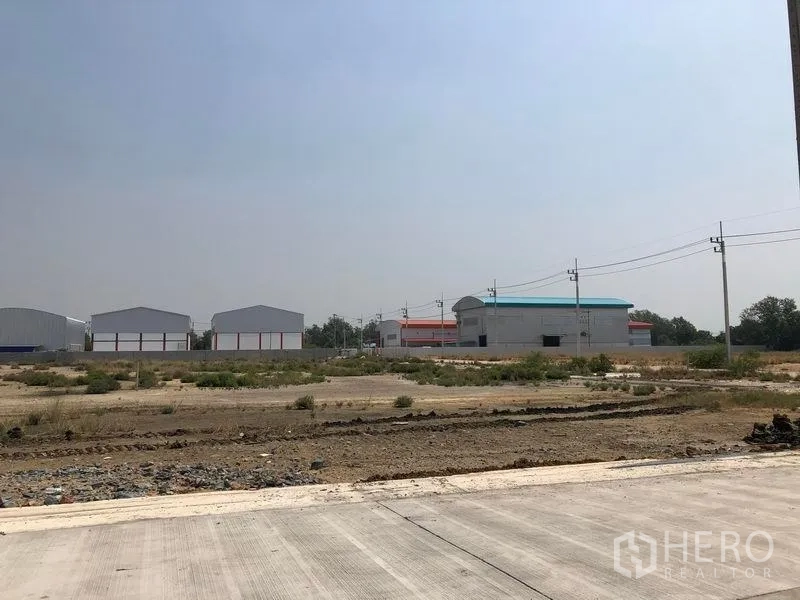 地 出售在Mueang Samut Sakhon, Samut Sakhon - 空地工业用地，前方混凝土路与电线杆，远处可见仓库厂房