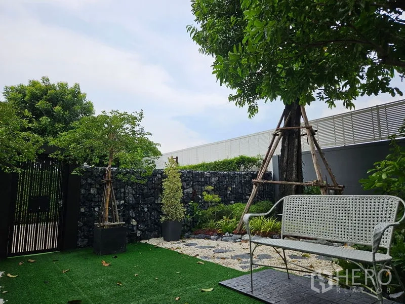Detached House for sale in Thawi Watthana, Bangkok - Front garden with artificial turf, bench and trees beside a stone wall gate.