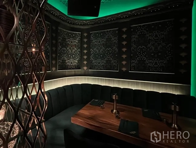零售空间 出租在Bang Rak, Bangkok - 氛围感私密卡座，弧形天鹅绒沙发、木桌与图案墙面，配绿色灯光。