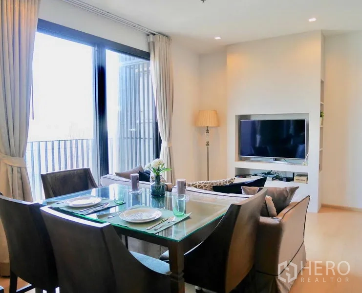 公寓 出租在Watthana, Bangkok - 餐区配玻璃餐桌与座椅，旁有电视墙与通往阳台的推拉门。