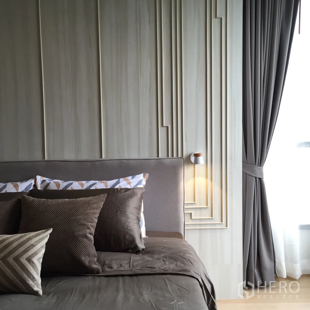 Condominium for rent or sale in Watthana, Bangkok - Cozy master bedroom with paneled feature wall, taupe bedding, bedside sconces and a sheer‑curtained window.