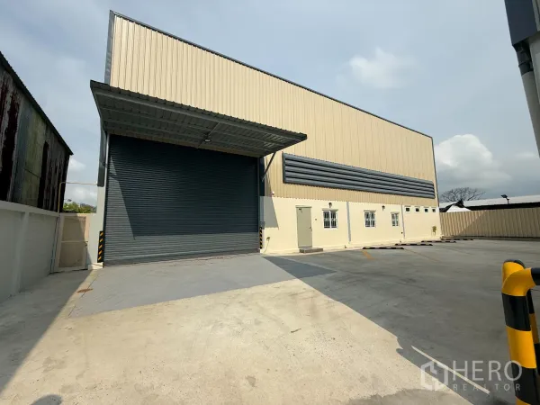 货栈 出租在Bang Khen, Bangkok - 曼谷邦肯区米色仓库外观，超大卷闸门、带顶装卸区及宽阔混凝土停车场。