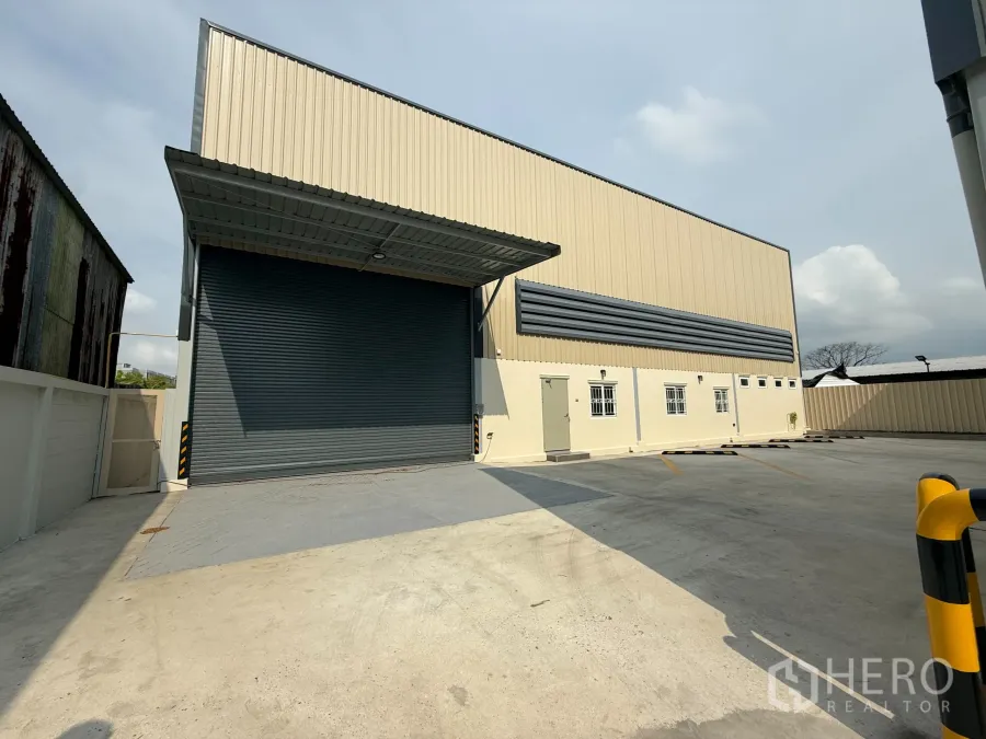 货栈 出租在Bang Khen, Bangkok - 曼谷邦肯区米色仓库外观，超大卷闸门、带顶装卸区及宽阔混凝土停车场。