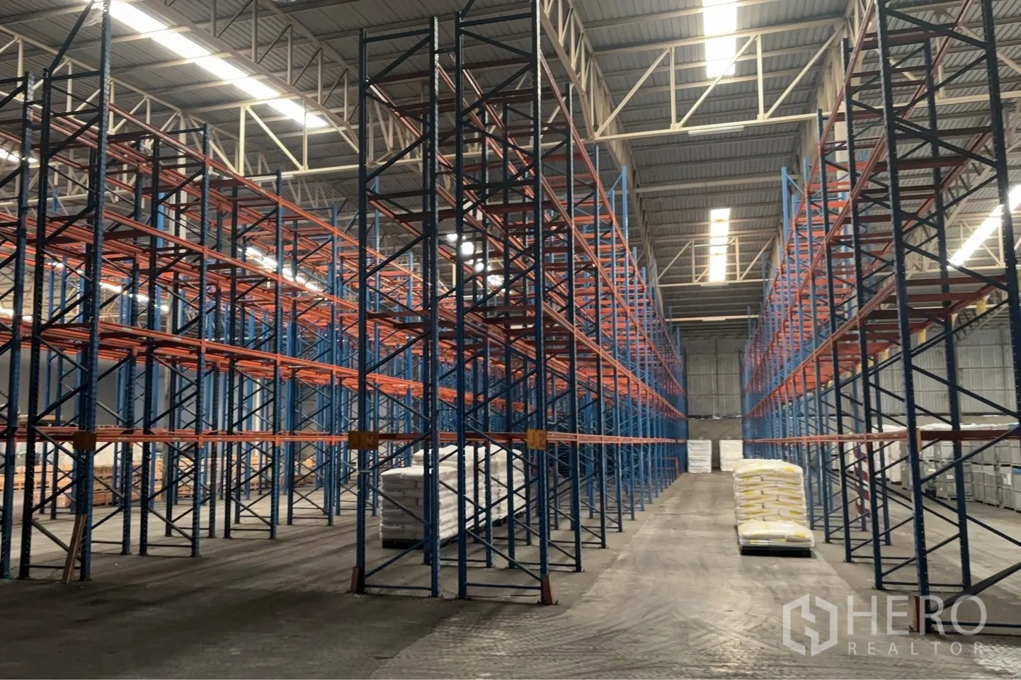 โกดัง for rent in Bang Pa-in, Phra Nakhon Si Ayutthaya - ทางเดินกว้างในโกดังเรียงรายด้วยชั้นวางสูง มีสินค้าแบบถุงวางบนพาเลทใต้ไฟส่องสว่าง