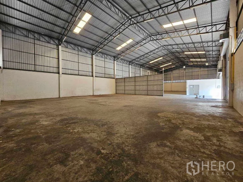 Warehouse for rent in Bang Bua Thong, Nonthaburi - Large storage hall with steel trusses, skylights, and a partitioned office area at the rear.