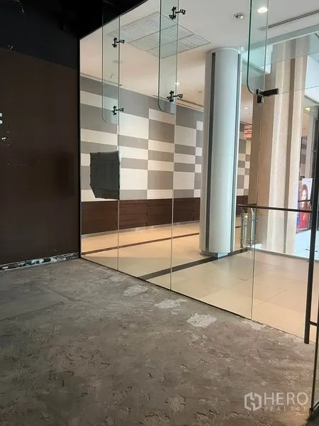 零售空间 出租在Watthana, Bangkok - 曼谷瓦他纳区购物走廊旁的小型商铺，落地玻璃门面与水泥地面，空间通透明亮。
