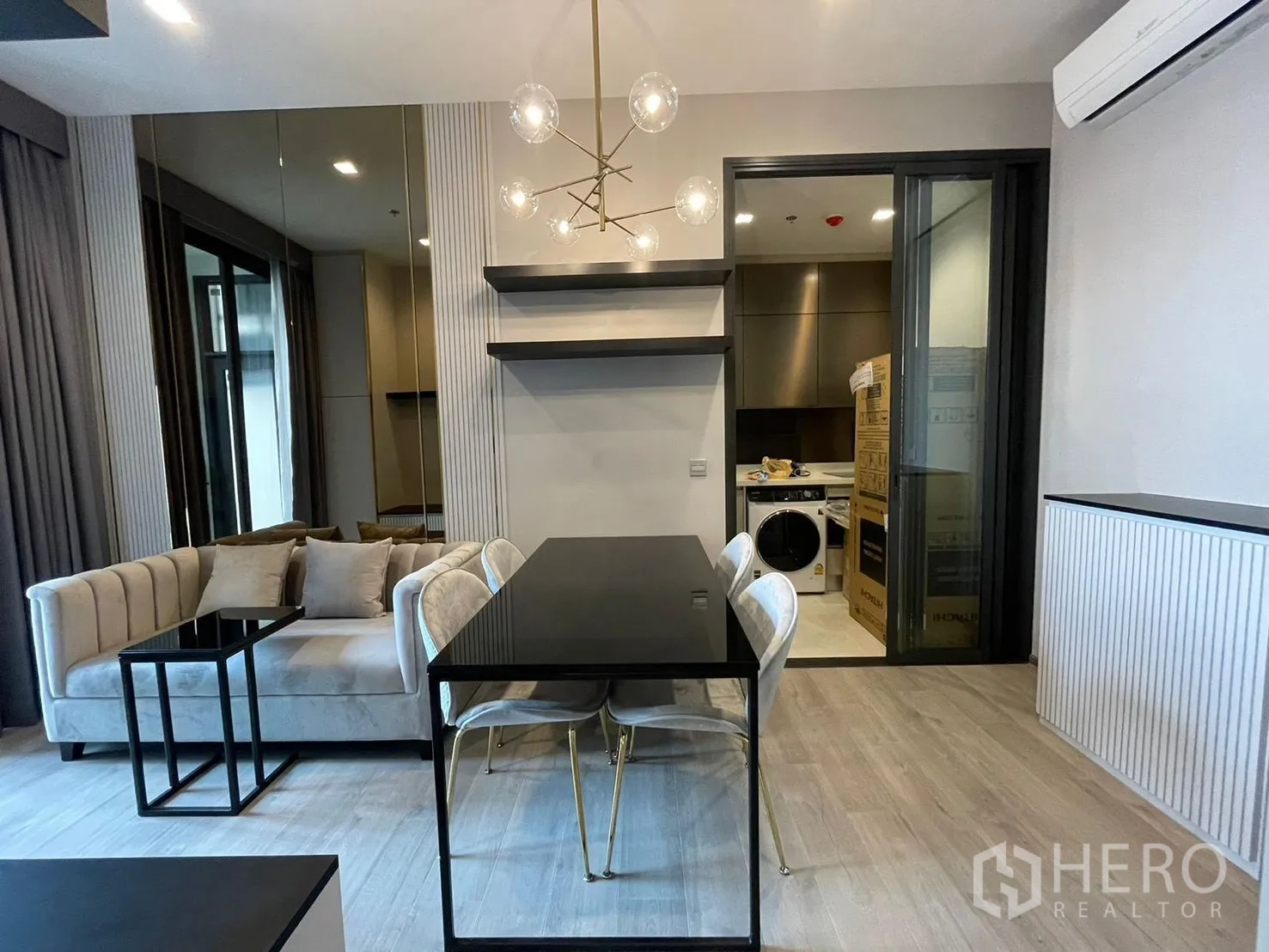 Condominium for rent in Ratchathewi, Bangkok - Contemporary living and dining space with plush sofa, black dining table, globe chandelier, and a sliding glass door to a separate kitchen with washing machine.